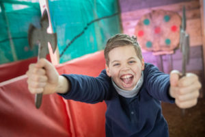 A young boy waves and smiles at the camera. He's holding two axes in his hands ready for axe throwing at Shrewsbury Prison