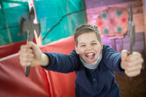A young boy waves and smiles at the camera. He's holding two axes in his hands ready for axe throwing at Shrewsbury Prison